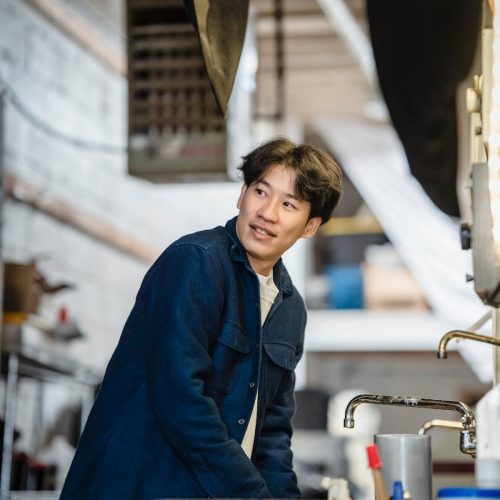 Man in a workshop wearing a blue jacket, engaged in industrial work.